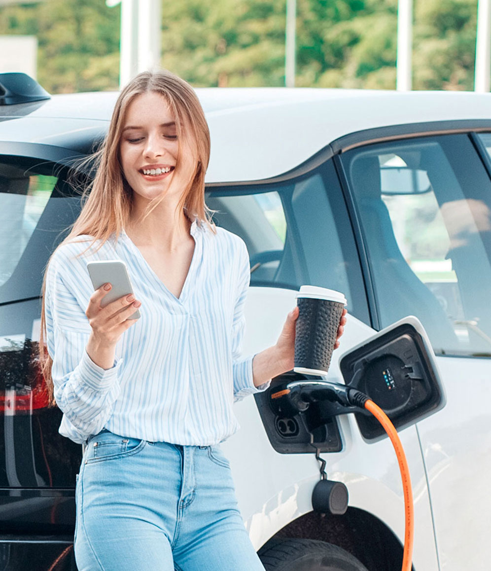 Frau schaut auf ihr Handy und hält einen Kaffeebecher in der Hand während ihr Elektroauto neben Ihr an einer Ladesäule lädt.