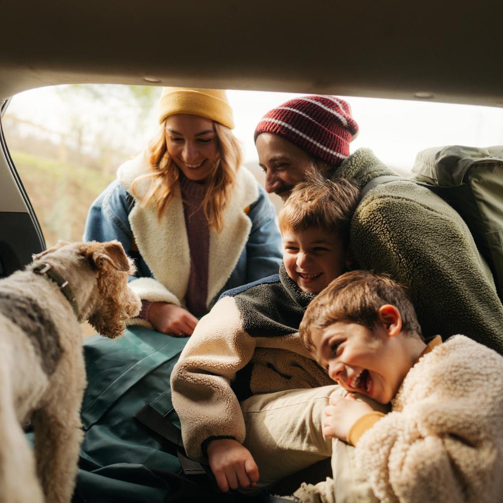Eine lächelnde, fröhliche Familie packt im Kofferraum ihres Autos ihre Sachen für ein Herbstabenteuer mit ihrem Hund