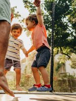 Der Vater springt mit seinen beiden Kindern an der Hand auf dem Trampolin im Garten.