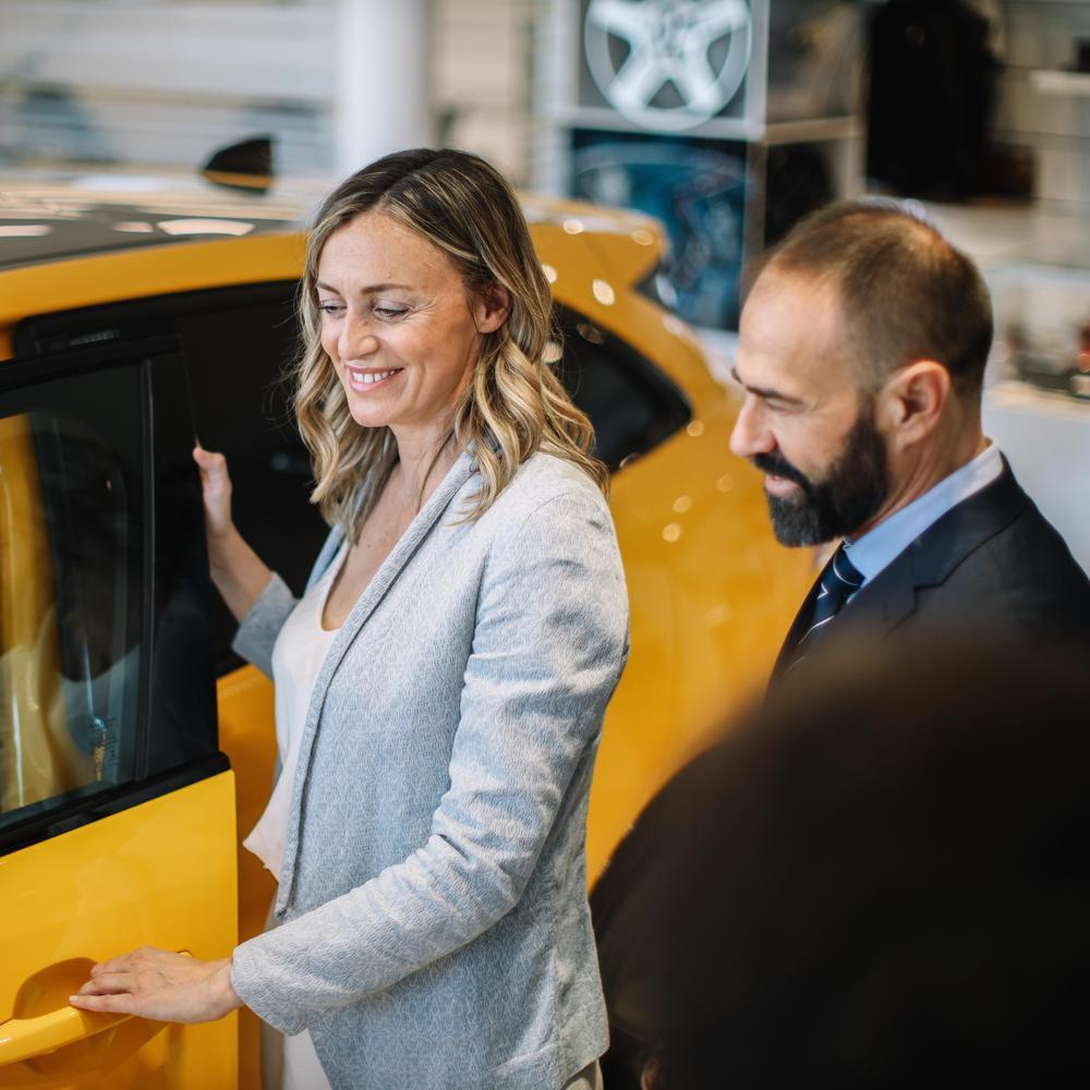 Frau steht mit einem Berater im Autohaus und öffnet die Tür eines gelben Autos