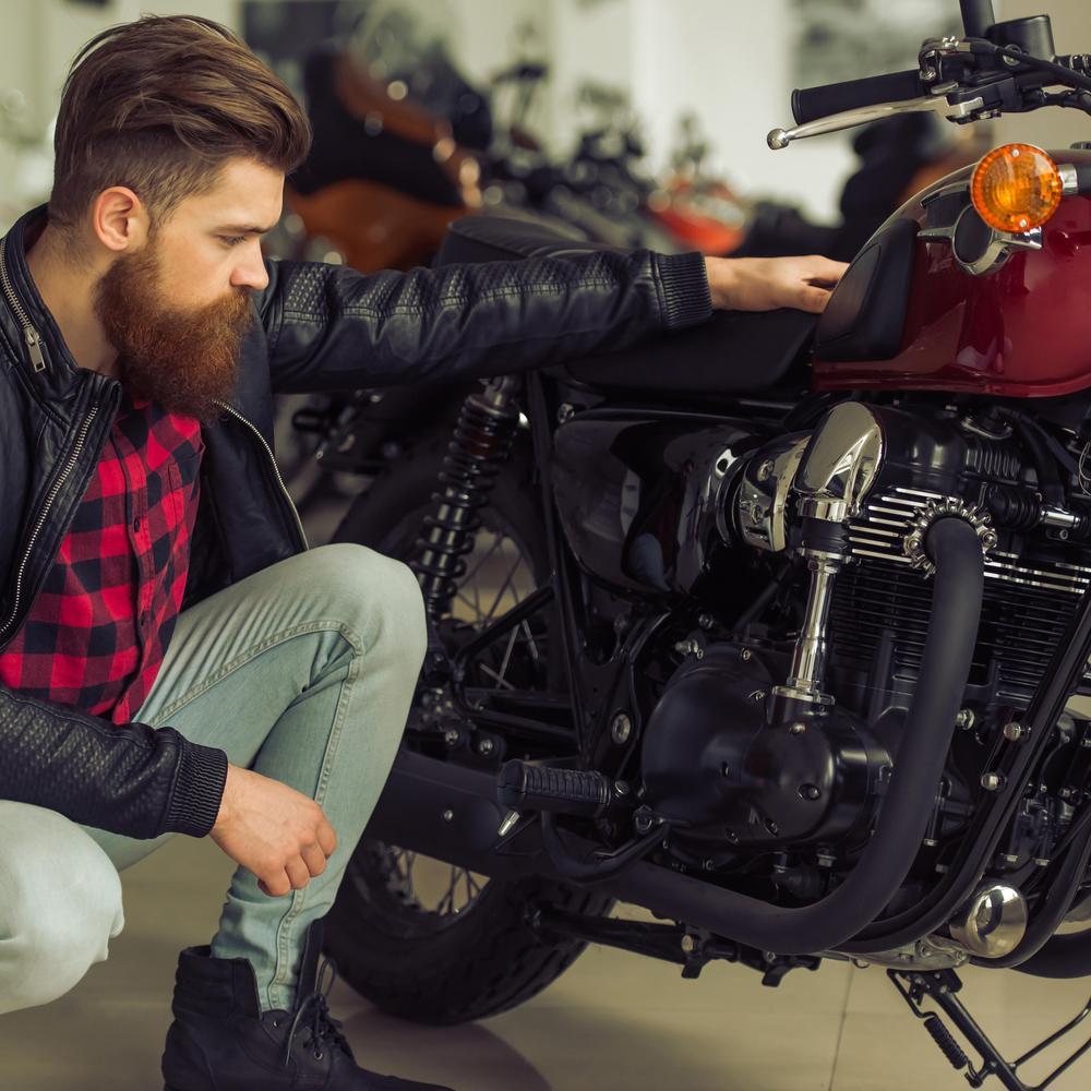 Ein  Mann mit Bart in schwarzer Lederjacke schaut sein Motorrad an.