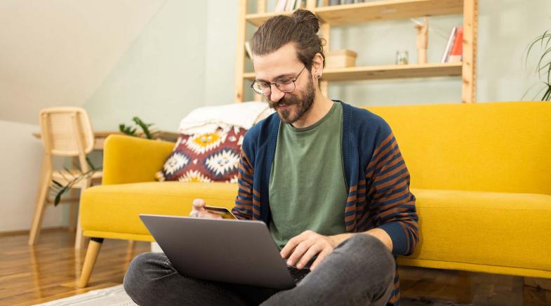 Ein junger Mann sitzt in seinem Wohnzimmer auf dem Boden und hat sein Laptop auf dem Schoß.
