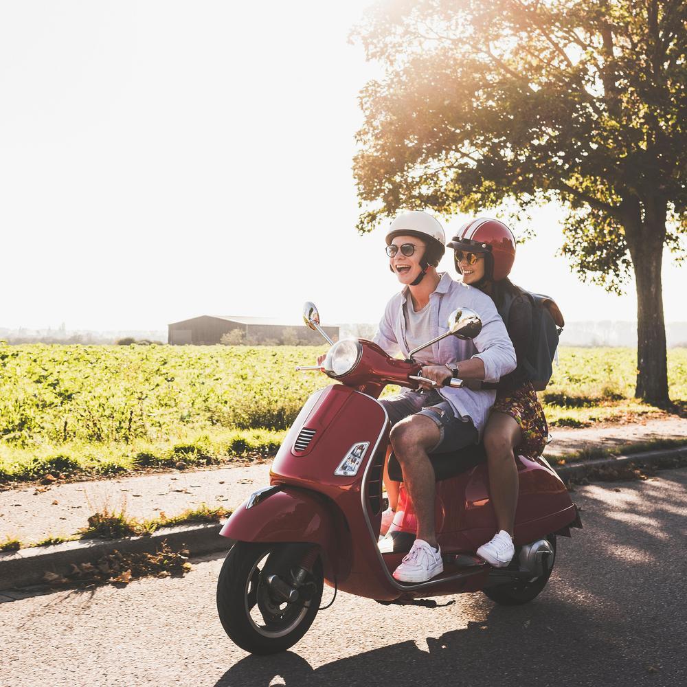 Glückliches junges Paar fährt im Sommer mit  Motorroller auf Landstraße.