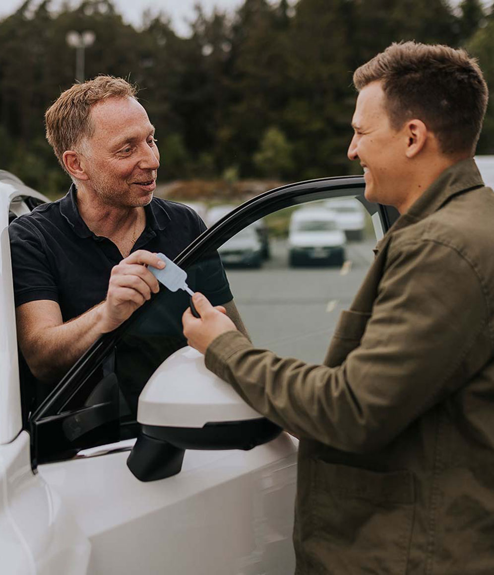 Zwei Personen stehen neben einem Auto auf einem Parkplatz. Eine Person übergibt der anderen eine Karte.