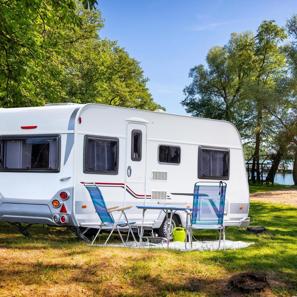 Idyllische Sommerszene am See - ein Wohnwagen und Campingmöbel stehen am Ufer zwischen Bäumen.