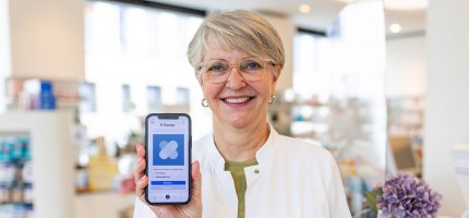 Eine Person in einem weißen Kittel zeigt ein Smartphone mit einer E-Rezept-App in einer Apotheke.