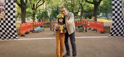 Herr Möller hält in der Zieleinfahrt einer Seifenkistenrennstrecke einen kleinen Jungen mit Helm im Arm, der einen Pokal gewonnen hat und reckt den Daumen nach oben.