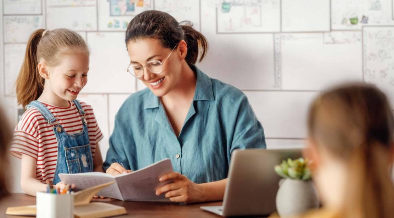 Eine Lehrerin sitzt in der Schule an ihrem Schreibtisch und erklärt ihrer Schülerin etwas in einem Heft.