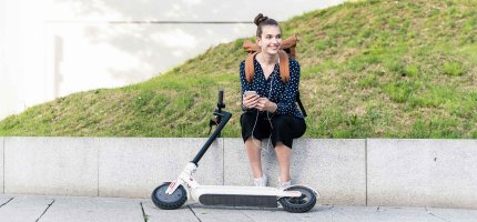 Junge Frau sitzt auf einer niedrigen Mauer. Vor ihr liegt ein E-Scooter.