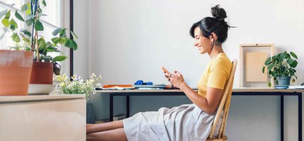 Frau sitzt am Schreibtisch und hat die Beine auf einem Hocker hochgelegt. Dabei schaut sie auf ihr Handy.