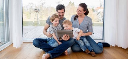 Eine junge Familie sitzt mit zwei Kindern im Haus auf einem Parkettboden und schaut in ein Tablett.