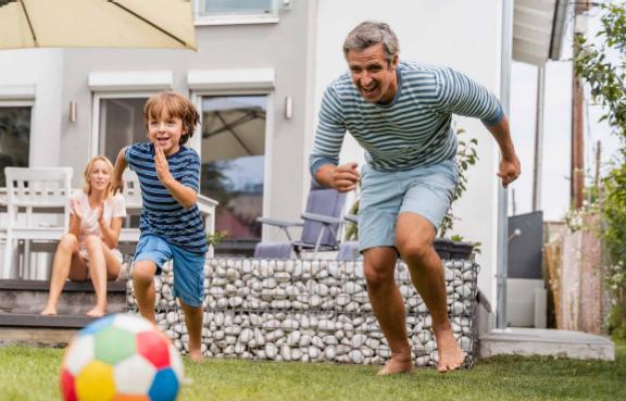 Ein Vater spielt mit seinem Sohn im Garten Fußball während die Mutter auf der Terrasse sitzt und zuschaut.