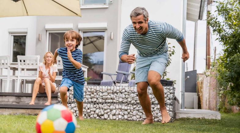 Ein Vater spielt mit seinem Sohn im Garten Fußball während die Mutter auf der Terrasse sitzt und zuschaut.