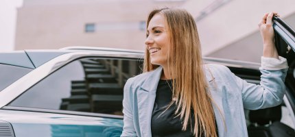 Junge Frau steigt aus dem Auto und hält ihr Smartphone locker in der Hand.