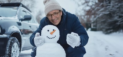 Herr Möller sitzt in einer verschneiten Einfahrt. Neben seinem Auto steht ein Schneemann.