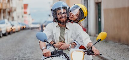 Pärchen fährt fröhlich auf einem Moped über eine Pflasterstraße.