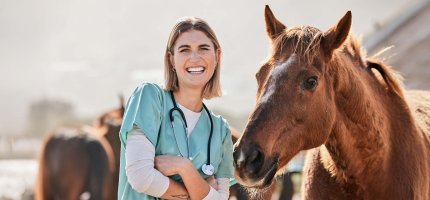 Eine junge Tierärztin steht neben einem Pferd und lacht in die Kamera.