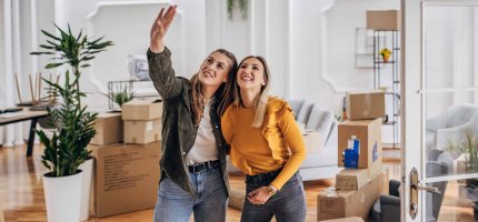 Zwei junge Frauen stehen zusammen und betrachten Ihre neue Wohnung voller Umzugskartons.