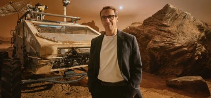 Herr Möller steht vor einem staubigen, futuristischen Geländerover in einer rötlichen Marslandschaft mit großen Felsen und einem hellen Himmelskörper am Himmel. Herr Möller trägt ein dunkles Sakko über einem weißen Shirt.