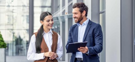 Mann und Frau in Business-Kleidung schauen außerhalb eines Bürogebäudes auf ein Tablet.