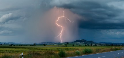 Blitz zuckt aus einer dunklen Unwetterwolke am Horizont neben einem Hügel in den Boden.