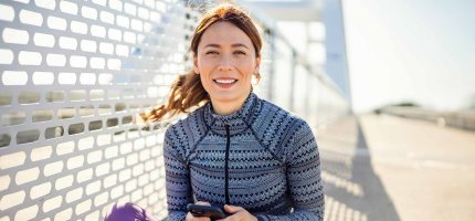 Eine Frau sitzt auf einer Brücke in Sportklamotten und hat ihr Handy in der Hand.