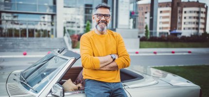 Älterer Mann lehnt in einer Großstadt an seinem Oldtimer-Cabrio