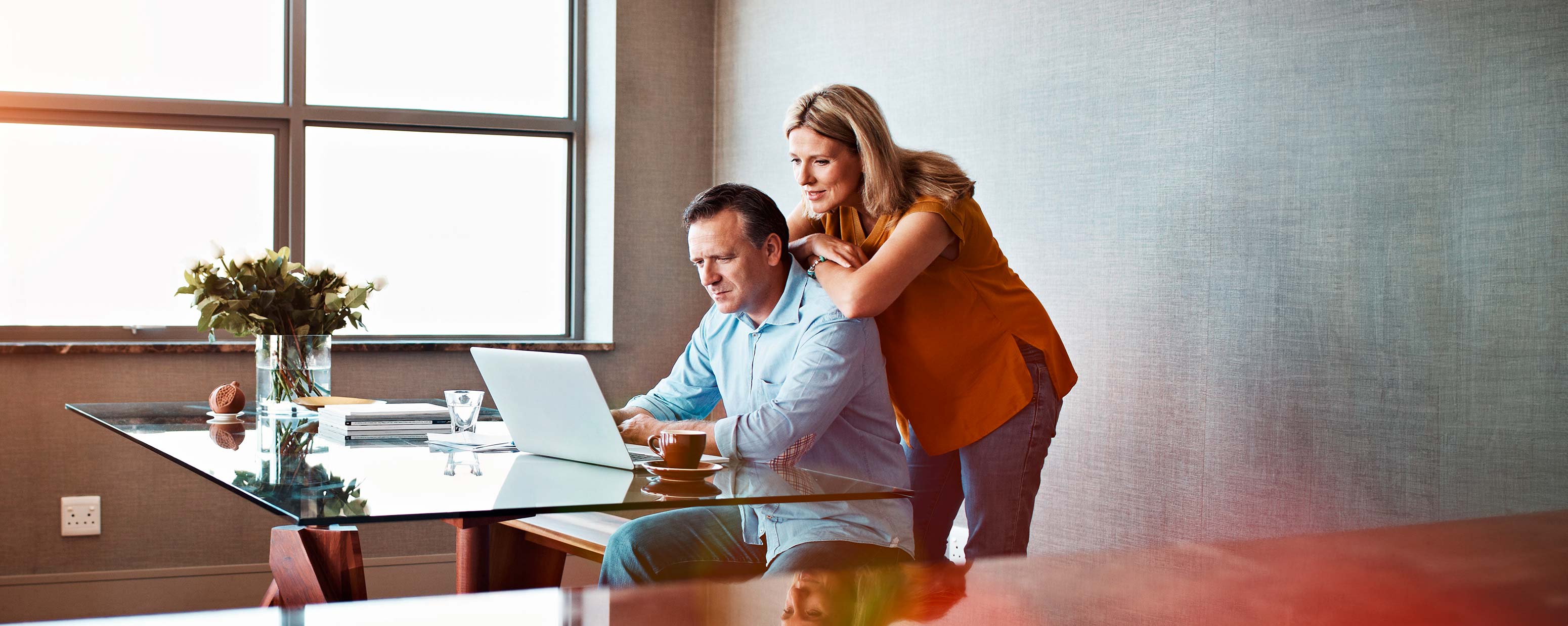 Älterer Mann sitz an einem gläsernen Schreibtisch und blick auf einen Laptop während sich eine Frau von hinten auf ihn stützt.