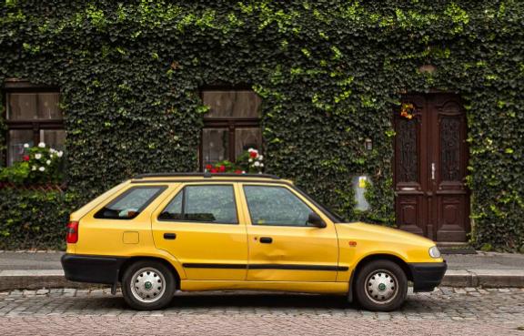 Ein alter Kombi mit Dellen steht am Straßenrand vor einem Haus, dessen Fassade mit Pflanzen bewachsen ist.