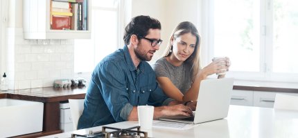 Paar sitzt in der Küche vor dem Laptop und schaut konzentriert.