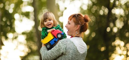 Eine Frau hält lachend ein Kleinkind hoch, das einen bunten Spielzeuglastwagen in der Hand hält.