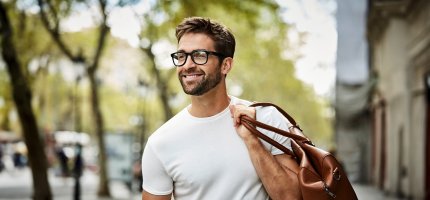 Mann mit Brille läuft mit einer Tasche über der Schulter lächelnd durch die Stadt.