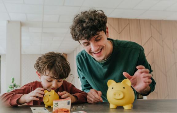 Vater und Sohn sitzen lächelnd am Tisch auf dem Geld und 2 Sparschweine liegen.
