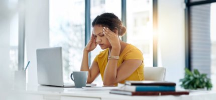 Frau sitzt mit geschlossenen Augen vor ihrem Laptop im Büro und hält sich mit beiden Händen die Schläfen.