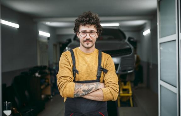 Ein tätowierter Automechaniker vor einer Hebebühne mit Auto.