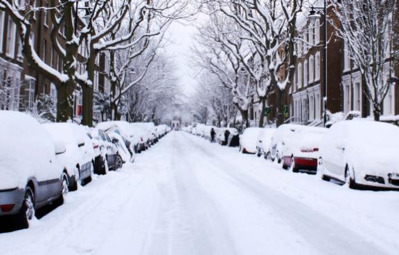 Verschneite Wohnstraße mit eingeschneiten parkenden Autos, kahlen Bäumen und Reihenhäusern links und rechts.