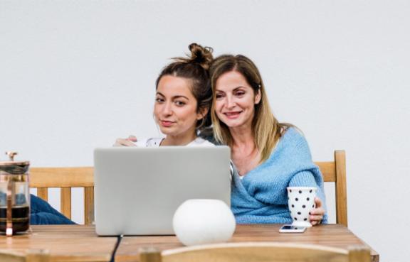 Mutter und Tochter sitzen am Tisch, trinken Kaffee und schauen auf einen Laptop.