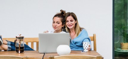 Mutter und Tochter sitzen am Tisch, trinken Kaffee und schauen auf einen Laptop.