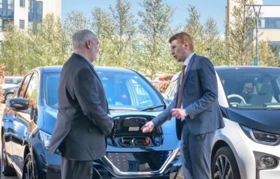 Autoverkäufer zeigt einem Kunden auf dem Parkplatz wo man ein E-Auto auflädt.