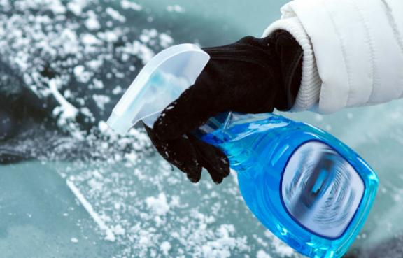 Eine Person sprüht mit einer blauen Flasche Enteisungsmittel auf eine vereiste Windschutzscheibe.