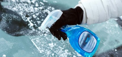 Eine Person sprüht mit einer blauen Flasche Enteisungsmittel auf eine vereiste Windschutzscheibe.