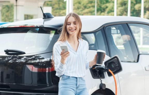Frau schaut auf ihr Handy und hält einen Kaffeebecher in der Hand während ihr Elektroauto neben Ihr an einer Ladesäule lädt.