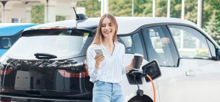 Frau schaut auf ihr Handy und hält einen Kaffeebecher in der hand während ihr Elektroauto neben Ihr an einer Ladesäule lädt.