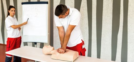 Eine Sanitäterin zeigt auf ein Flipchart und ein Sanitäter macht eine Herzdruckmassage an einer Erste-Hilfe-Puppe.