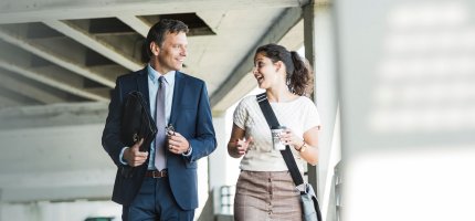 Ein Mann mit Anzug und Aktentasche und eine Frau mit Messenger-Bag und Reisekaffeebecher gehen nebeneinander und reden.