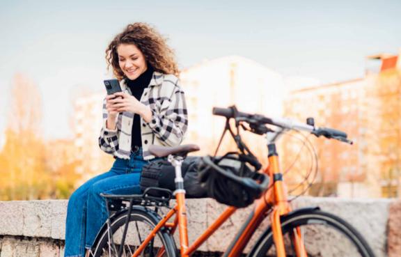 Frau sitzt auf einer Mauer und schaut auf Ihr Handy. Neben Ihr lehnt ihr Fahrrad an der Mauer.