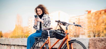 Frau sitzt auf einer Mauer und schaut auf Ihr Handy. Neben Ihr lehnt ihr Fahrrad an der Mauer.