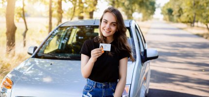 Junge Frau lehnt an ihrem Auto, das am Straßenrand einer Allee parkt, und zeigt ihren Führerschein.