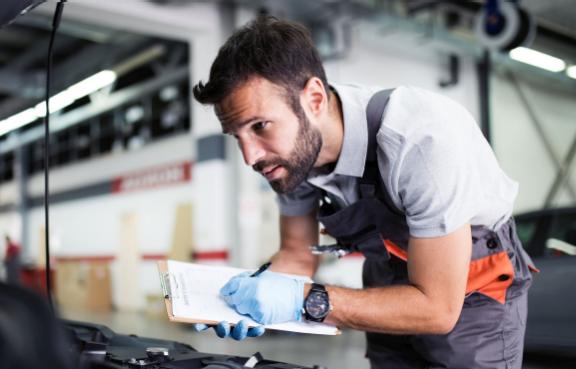 Ein Mechaniker geht in der Werkstatt eine Checkliste durch und schaut dabei in einen Motorraum eines Autos..