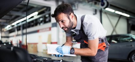 Ein Mechaniker geht in der Werkstatt eine Checkliste durch und schaut dabei in einen Motorraum eines Autos..